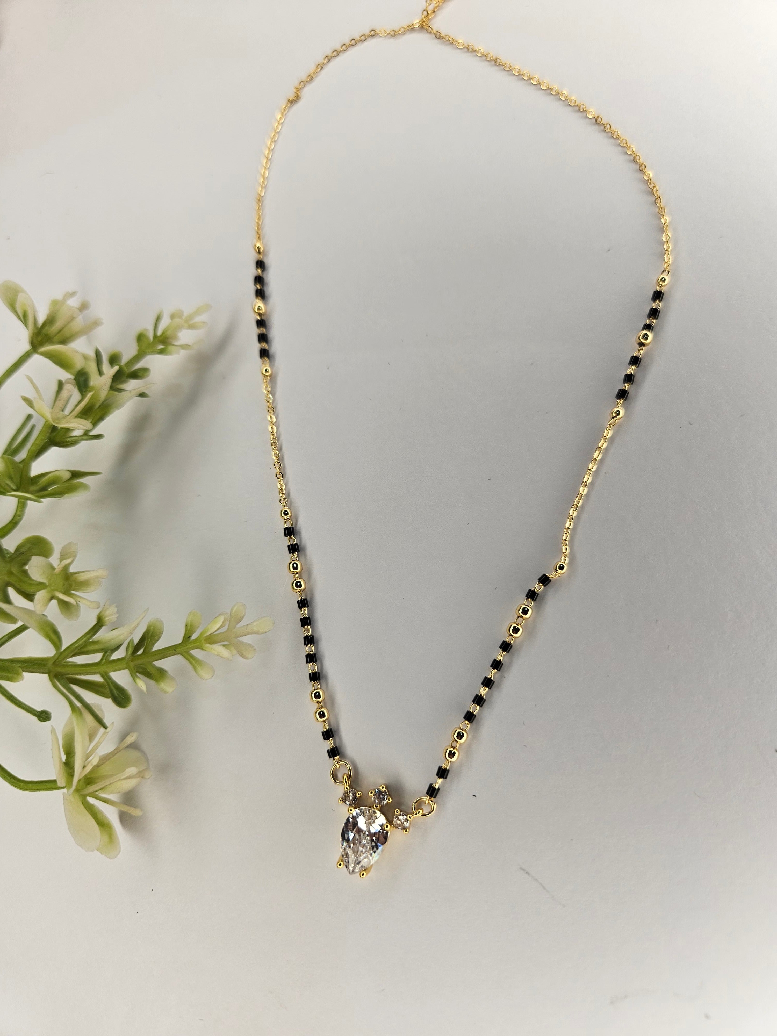A gold chain necklace with alternating black and gold beads, featuring a small gold pendant with a clear faceted gemstone, displayed on a light gray surface with decorative green leaves on the left.