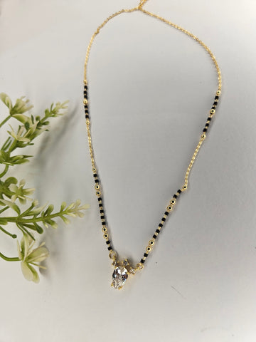 A gold chain necklace with alternating black and gold beads, featuring a small gold pendant with a clear faceted gemstone, displayed on a light gray surface with decorative green leaves on the left.