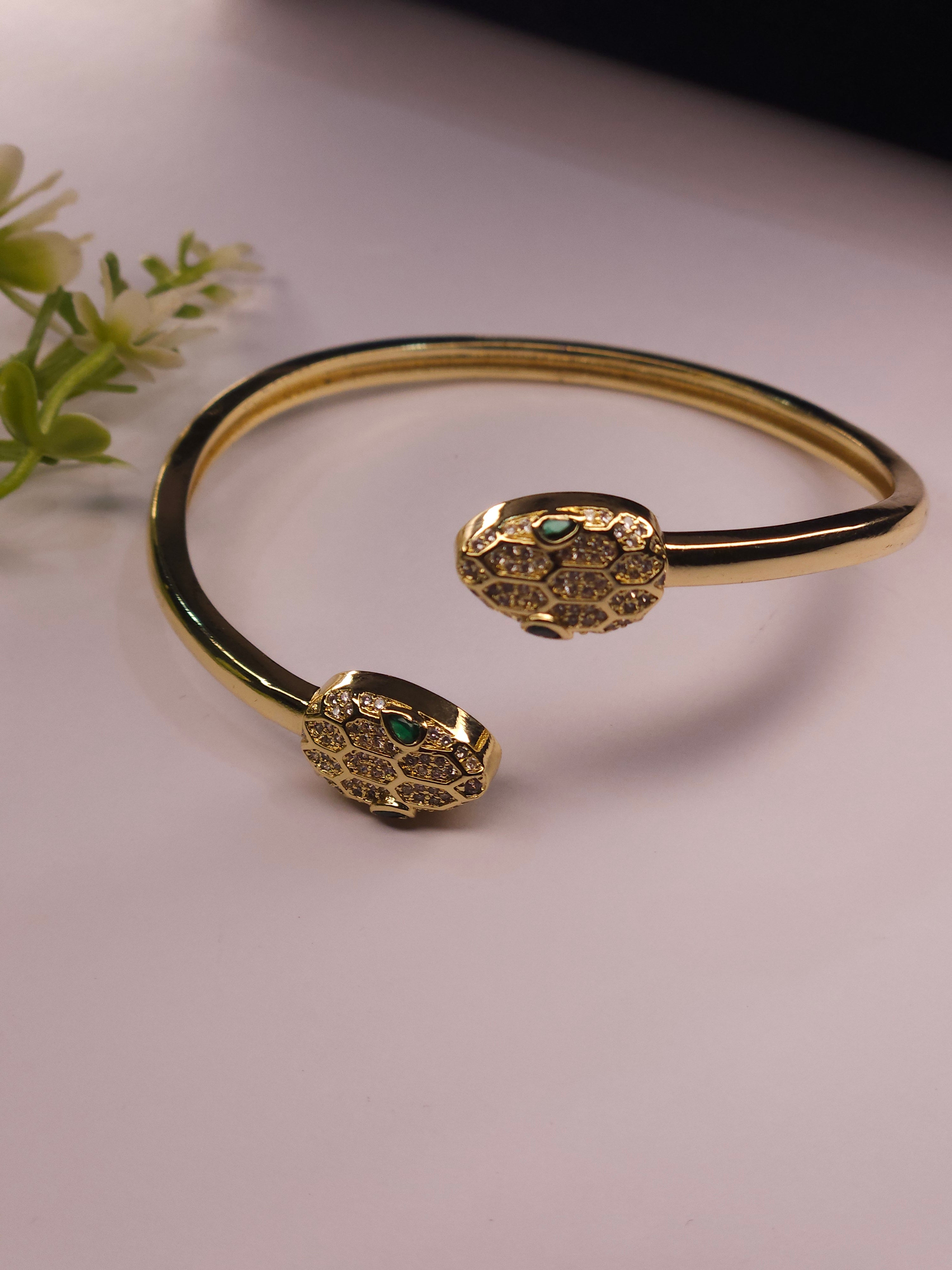 Gold snake-head cuff bracelet with small green stone eyes, photographed on a pale pink surface with a small floral prop in the corner.