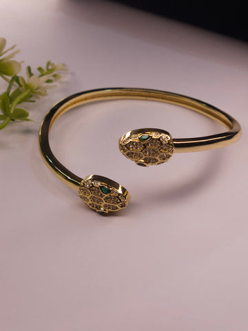 Gold snake-head cuff bracelet with small green stone eyes, photographed on a pale pink surface with a small floral prop in the corner.